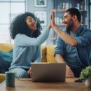 Diverse Multiethnic Couple are Sitting on a Couch Sofa in Stylish Living Room and Choosing Items to Buy Online with Laptop Computer, Give High Five. Friends or Colleagues are Discuss Work Projects.